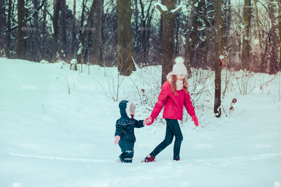 Teenage girl enjoying snow with her little sister. Children are walking through deep snow while snow falling, enjoying wintertime. Sisters spending time together. Girls are wearing winter clothes