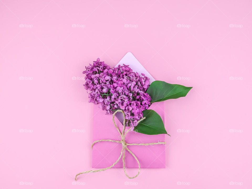 A branch of a lilac flower in an envelope tied with jute thread lies in the center on a pink background, flat lay close-up.