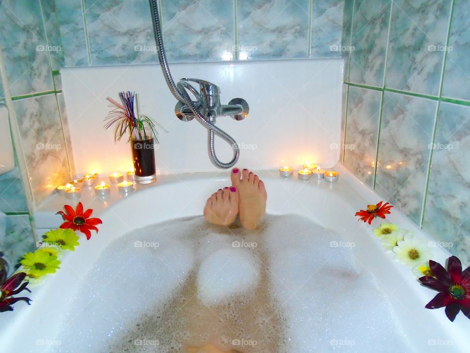 Woman's feet with red nail polish in bubble bath with candles and flowers