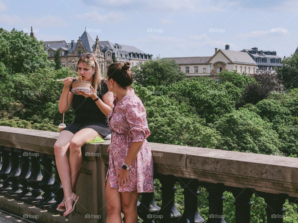 Two beautiful caucasian girls chatting among themselves sit on the railing of the bridge and eat Asian food with chopsticks on a summer day in Luxembourg city, close-up side view.