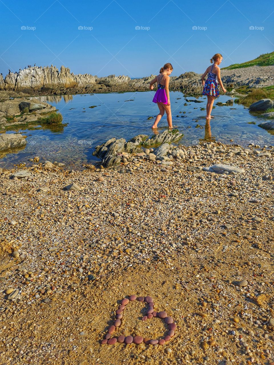 Two young sisters walking through a rock pool at the beach, enjoying the beautiful sunny day, with pepples arranged in a heart shape on the ground