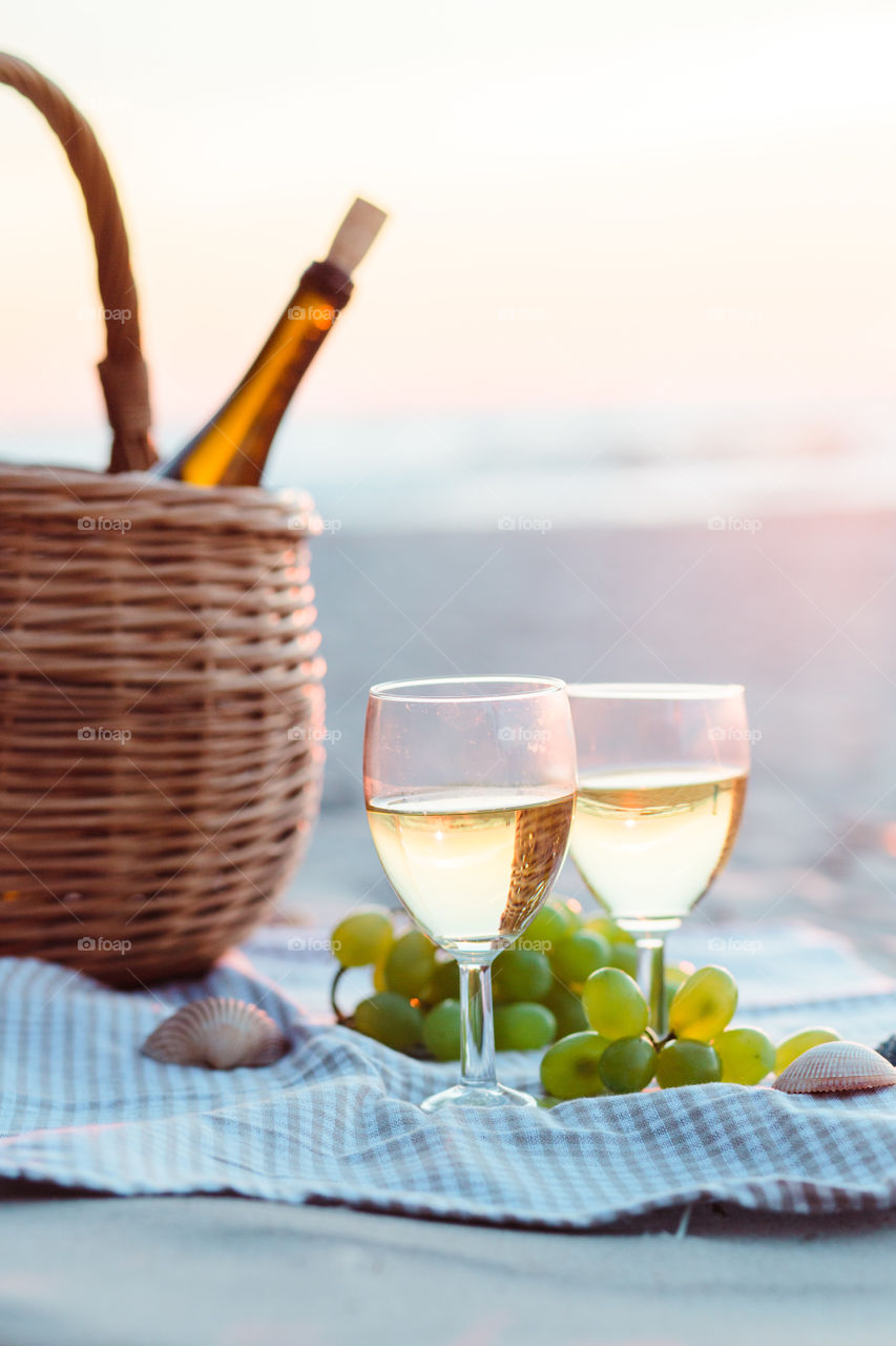 Two wine glasses with white wine standing on sand, on beach, beside grapes and wicker basket with bottle of wine. Sea waves in the background