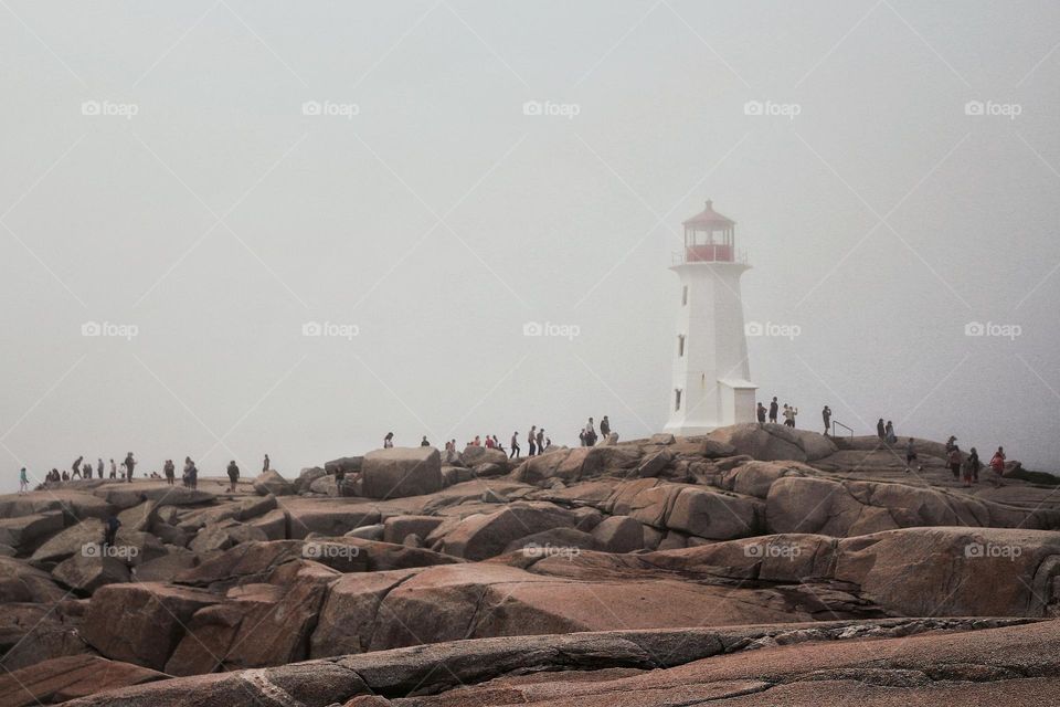 Crowds of tourists gathered on the rocks surrounding the Peggy's Cove lighthouse on a foggy afternoon.