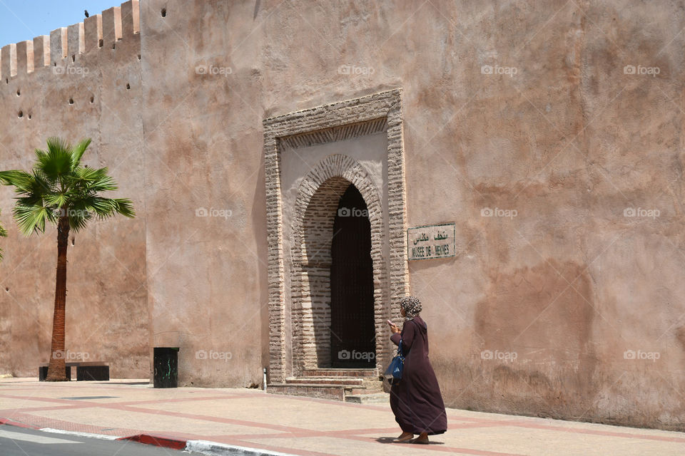 Moslim woman in long clothes and headscarf walking before a city castle wallwith a palm tree in Marocco