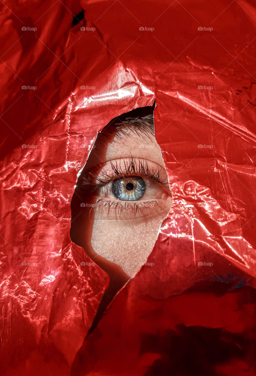 Close-up eye peeking through torn red paper.