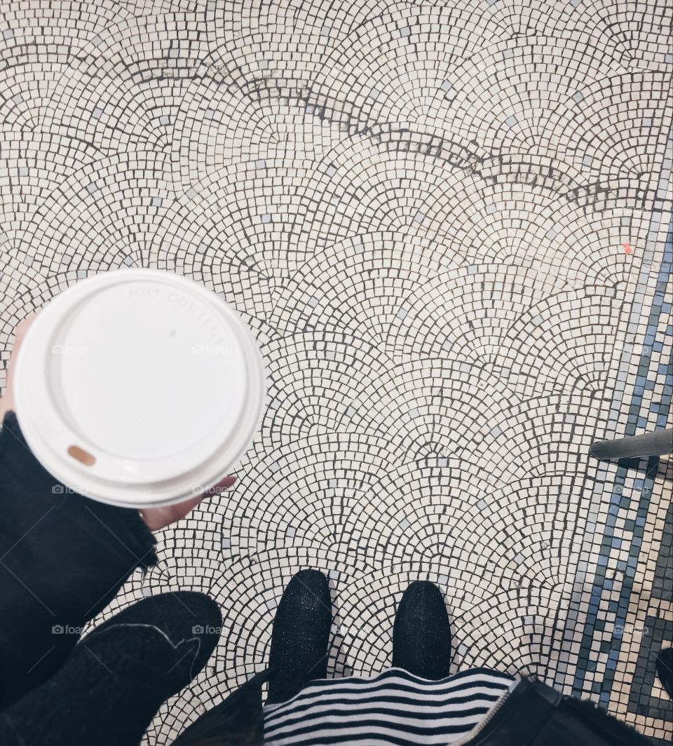 A female wearing black and white & black glittery shoes holds a Starbucks cup, looking town at the tiled floor below her