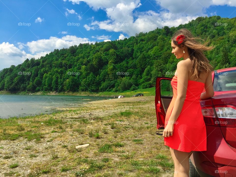 Woman in red dress standing near red car surrounded by lake and green forest in summer
