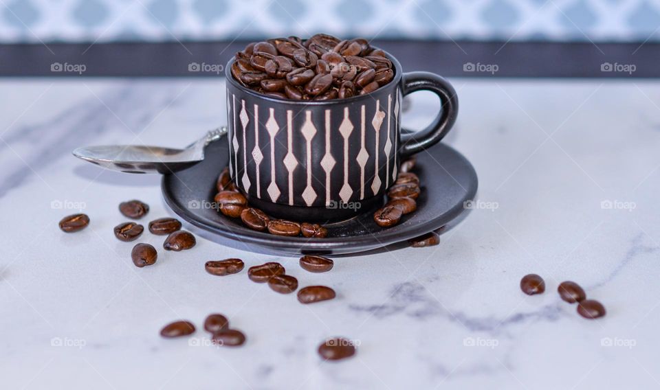 A tiny cup and saucer filled with coffee beans on a marble counter with a spoon