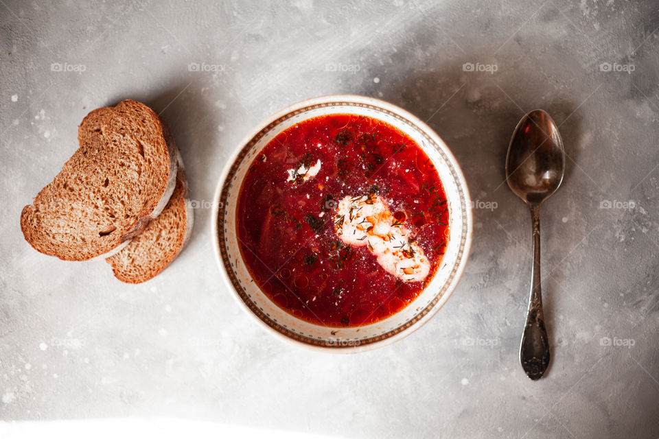 Plate of Ukrainian borsch. Nearby are two sliced pieces of bread and a spoon. A plate is on the newspaper. Photo taken on a white concrete background.Red borsch, add sour cream and greens.