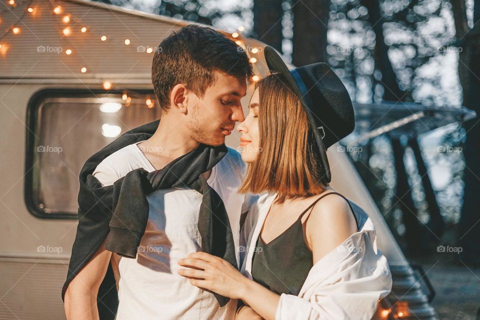 Young couple in love in stylish fashionable outfit romantic weekend wedding trip against background of camper in woods forest road trip