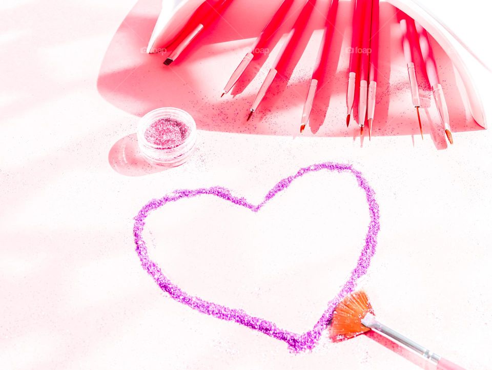 Lilac heart of nail glitter with pink brushes and nail drying lamp on a soft pink background, side view. Concept cosmetics and beauty salons.