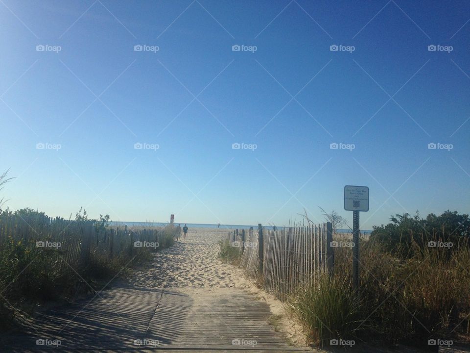 A view of a path to the beach in Cape May, NJ. People can be seen walking in the distance and a glimpse of the ocean is in the background.