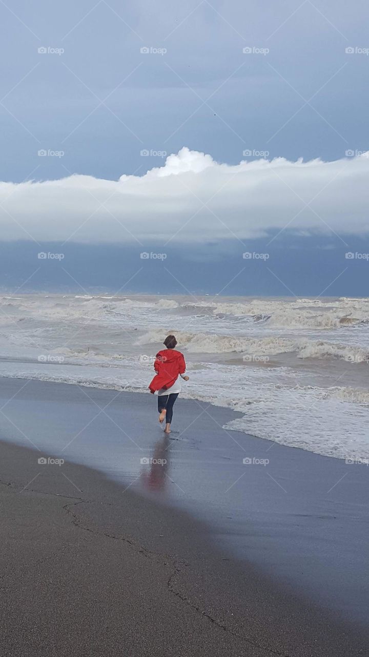 Waves, wind, fog and a girl in a red raincoat runs along the seashore during a storm.