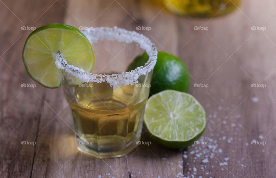 shot of tequila with salt and a lime wedge on the rim sitting on a wood table next to a sliced lime