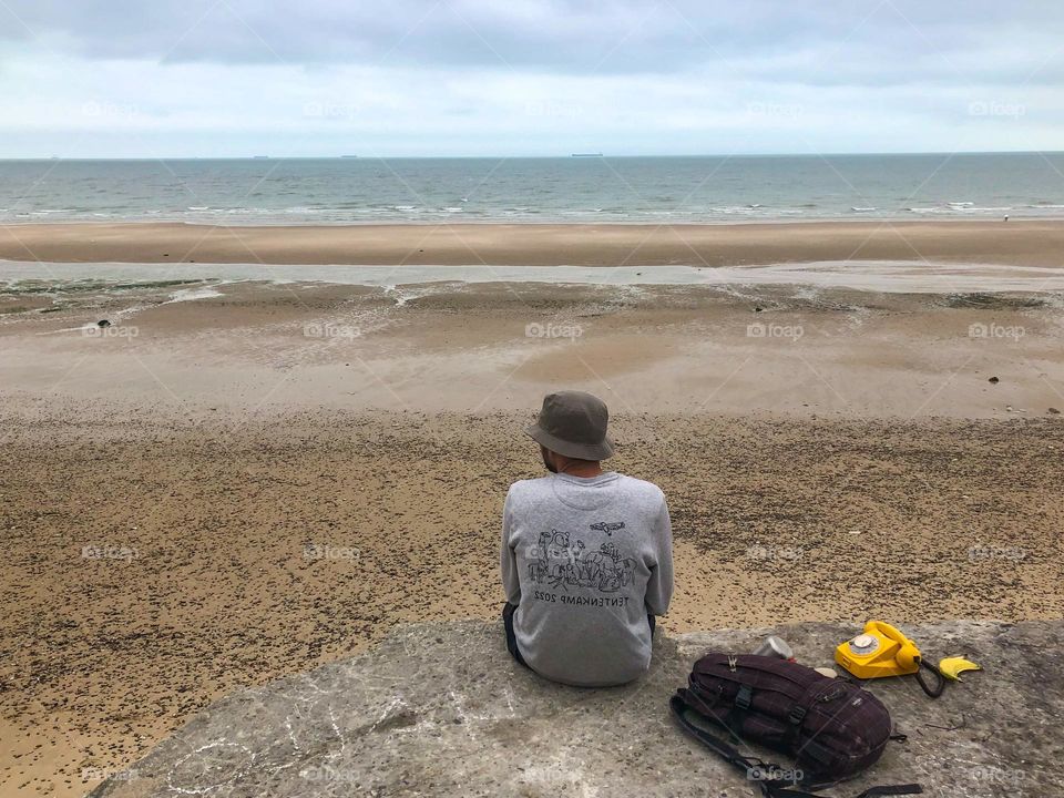 A man admiring the sea sitting on the beach with an old phone next to him. 