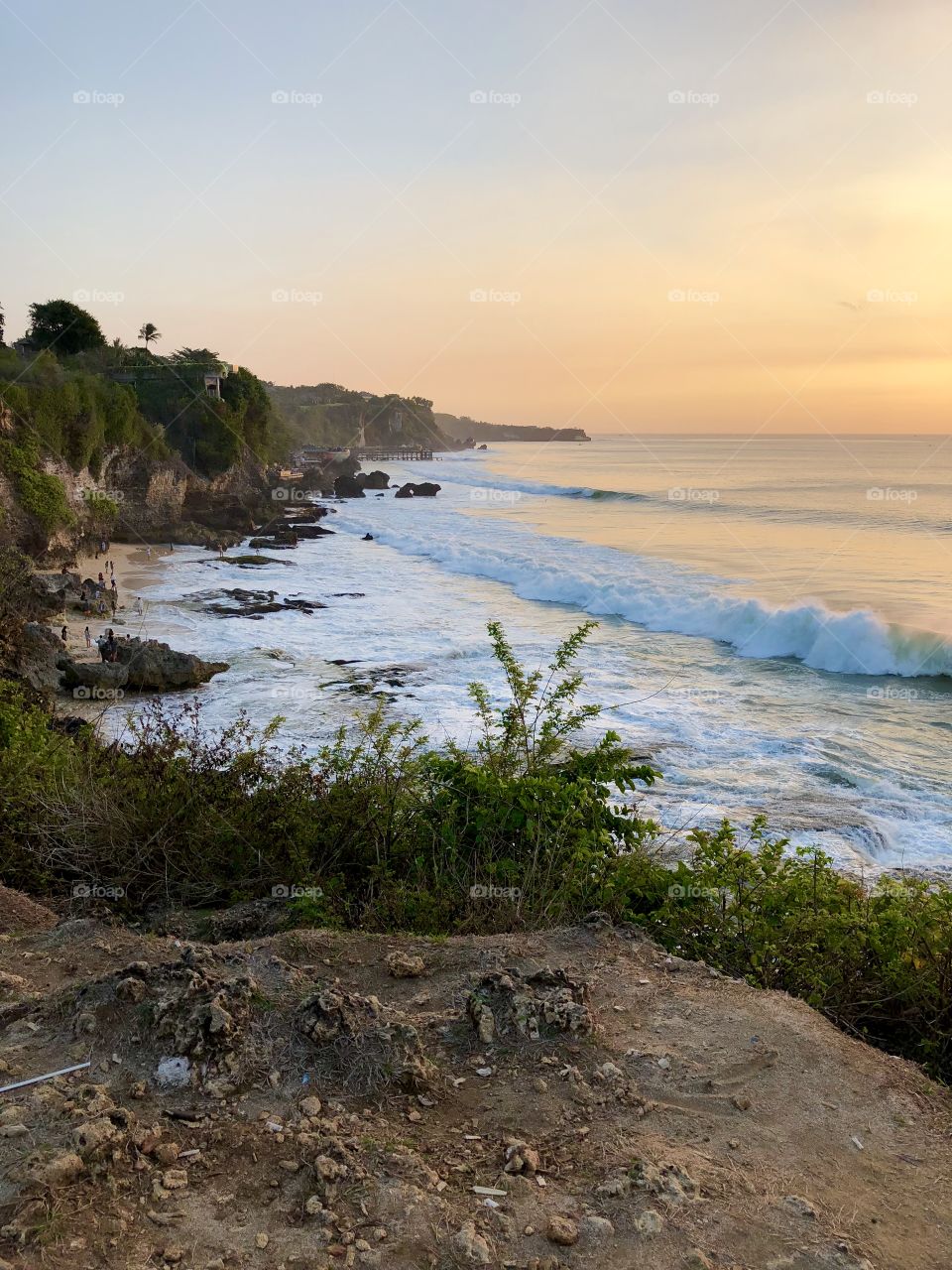 Sunset on curvy island coast. Big ocean waves. Dim lighting. Dirt in foreground. Developing country. Bali, Indonesia. 2018.