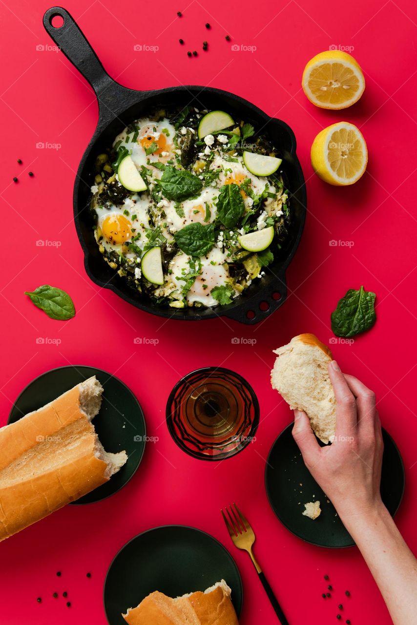 Flat lay of a green shakshuka dish in a cast iron skillet with a person’s hand reaching for a piece of bread 