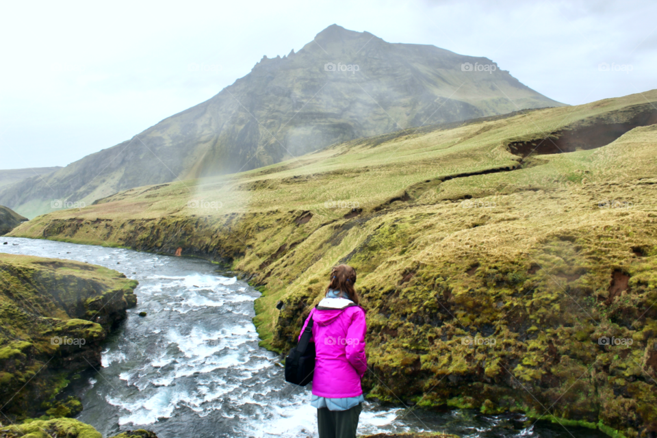 A woman overlooks the rushing river near a mountain and skogafoss waterfall in the Icelandic countryside