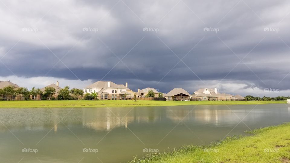 A neighborhood pond in front of houses, with dark storm clouds in the background.