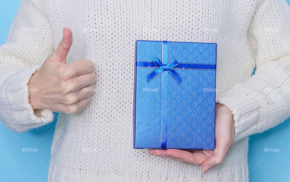 Young caucasian girl in a white knitted sweater holds a blue gift box with one hand and shows a cool gesture with the other on a light blue background,close-up side view. Holiday gifts concept.