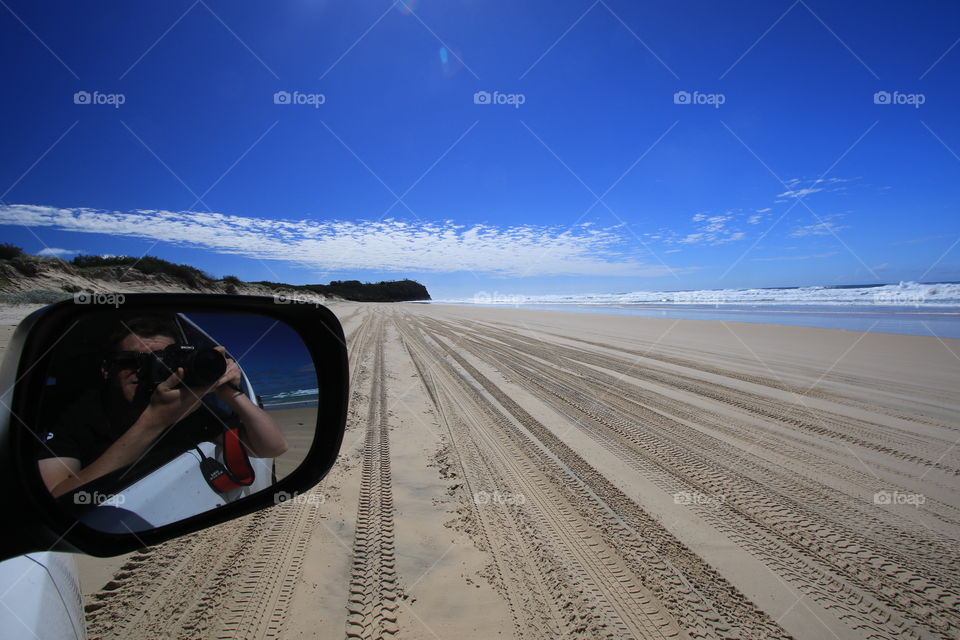 Fraser Island beach photo including photographer reflection in the car wing mirror. Car tracks in the sand.