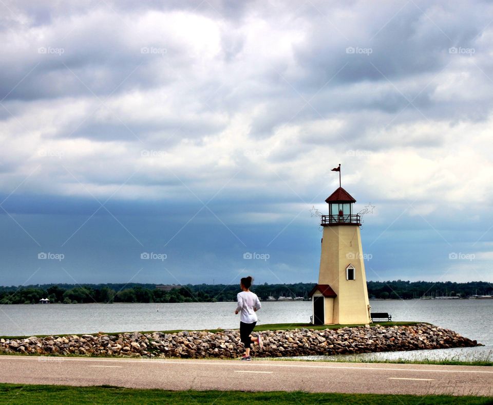 Lake Hefner in OKC with a lighthouse. A jogger is in the picture which represents the diverse use of the lake now. Since MAPS for OKC was implemented with a penny sales tax for specific projects, it has become a hotspot for restaurants, water sports, naturists, fishermen, and exercise enthusiasts.