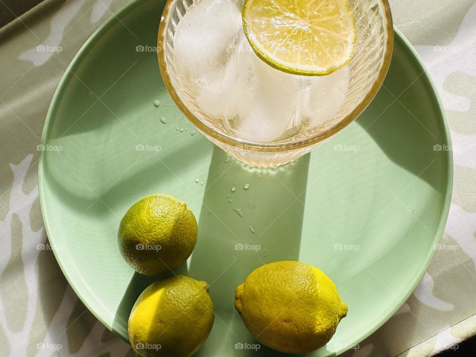 A glass of seltzer water with ice and a slice of lime, just picked from the tree, on a green plate sitting in warm afternoon sun and shadows