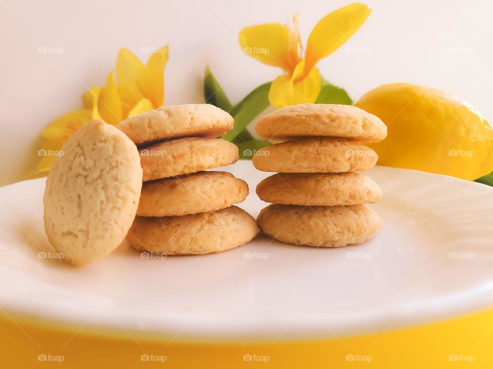 Nine lemon cookie snaps stacked on a white saucer on a yellow surface;  accented by yellow flowers and a lemon against a white background.