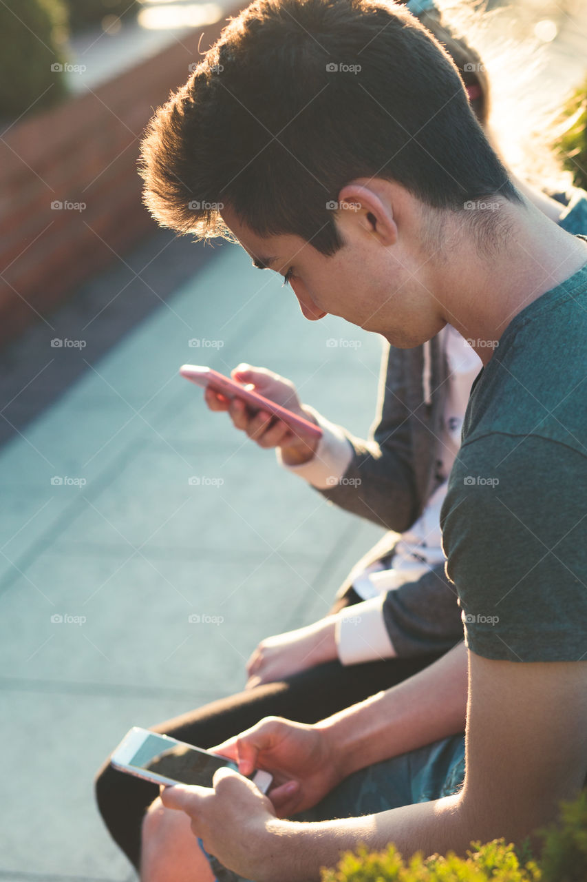 Couple of friends, teenage girl and boy, having fun together with smartphones, sitting in center of town, spending time together. Real people, authentic situations