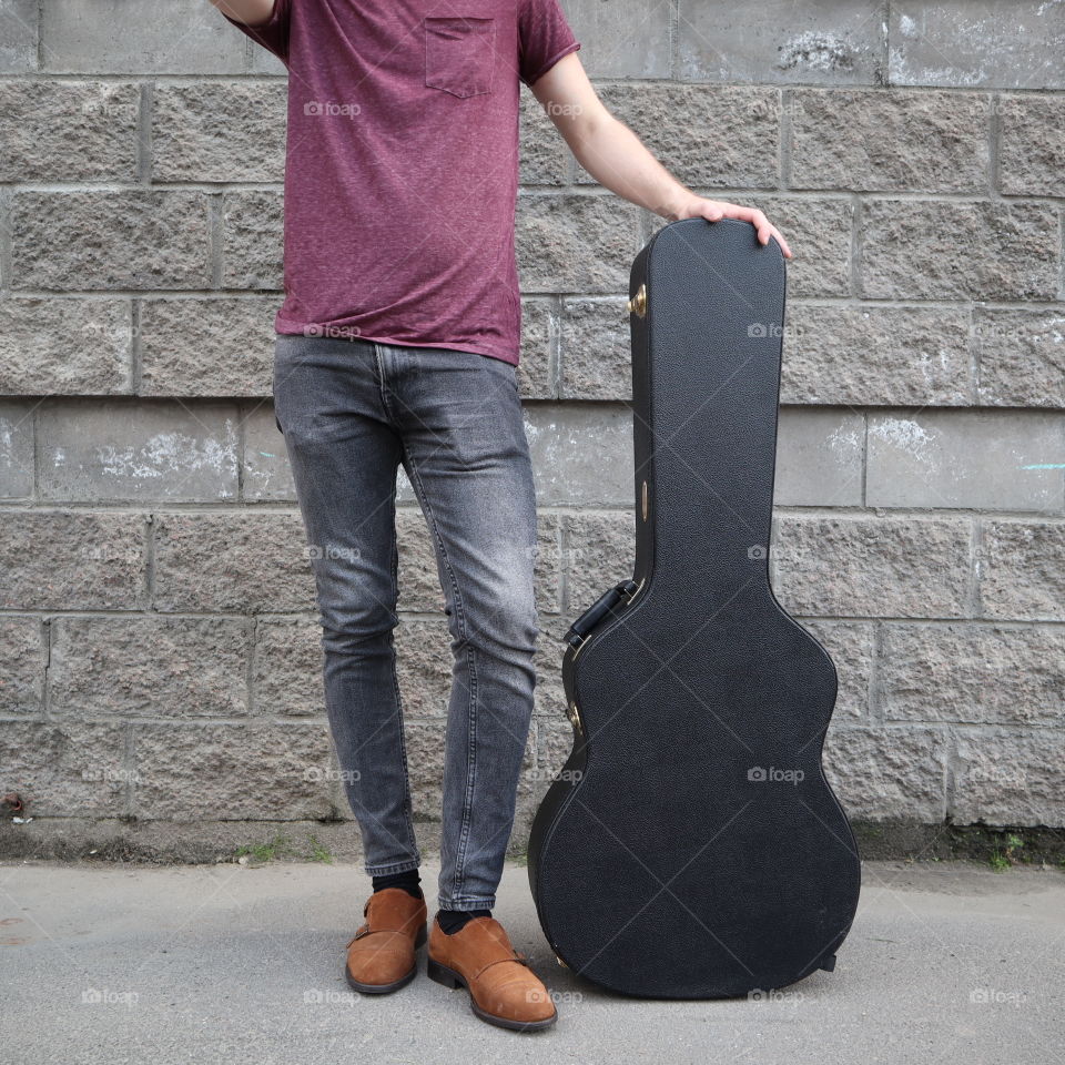 man put his foot on a hard guitar case. hard case for electric guitar. Man dressed in jeans holding guitar case against wall. guy with a guitar. copy space text