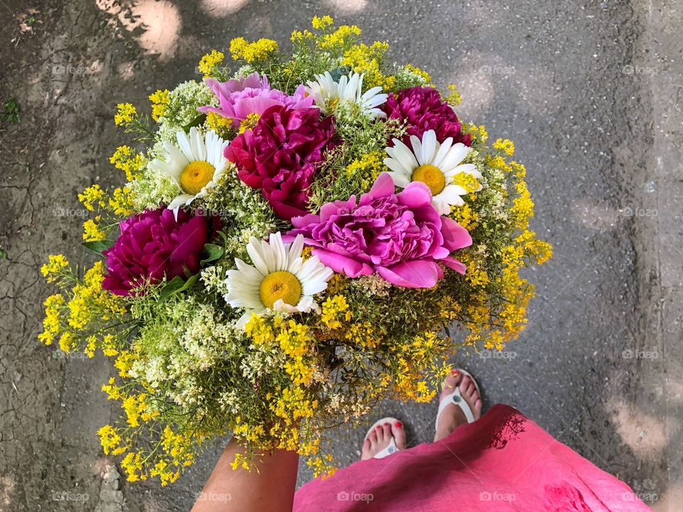 Woman in pink dress holding a giant bouquet of summer flowers consisting of pink peonies, daisies and yellow goldenrod