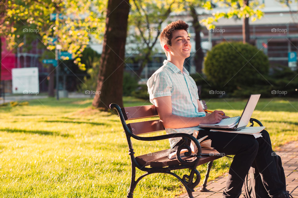 Student working on a laptop using books and notes sitting on a bench in a park. Young boy wearing a blue shirt and dark jeans