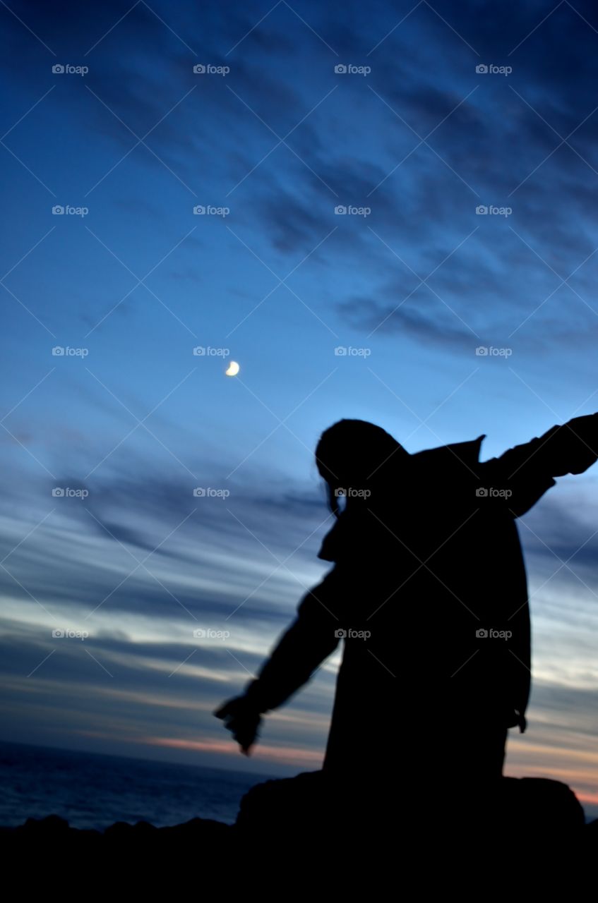 A girl’s silhouette showing the joy and that rewarding feeling after a hike as she stands beneath the crescent moon and cirrus clouds on a clear indigo night sky. 