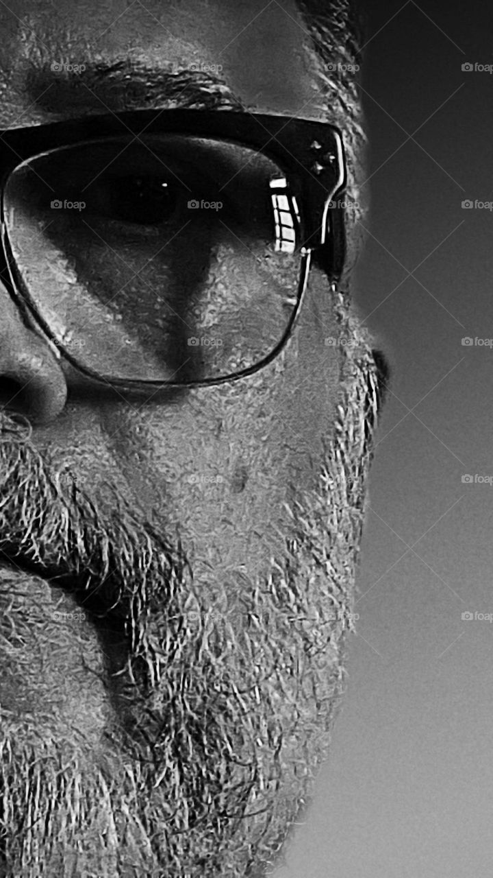 Half face of a man with a beard and glasses.  Reflection of part of the window in the eyeglass lens and shadows on the face.