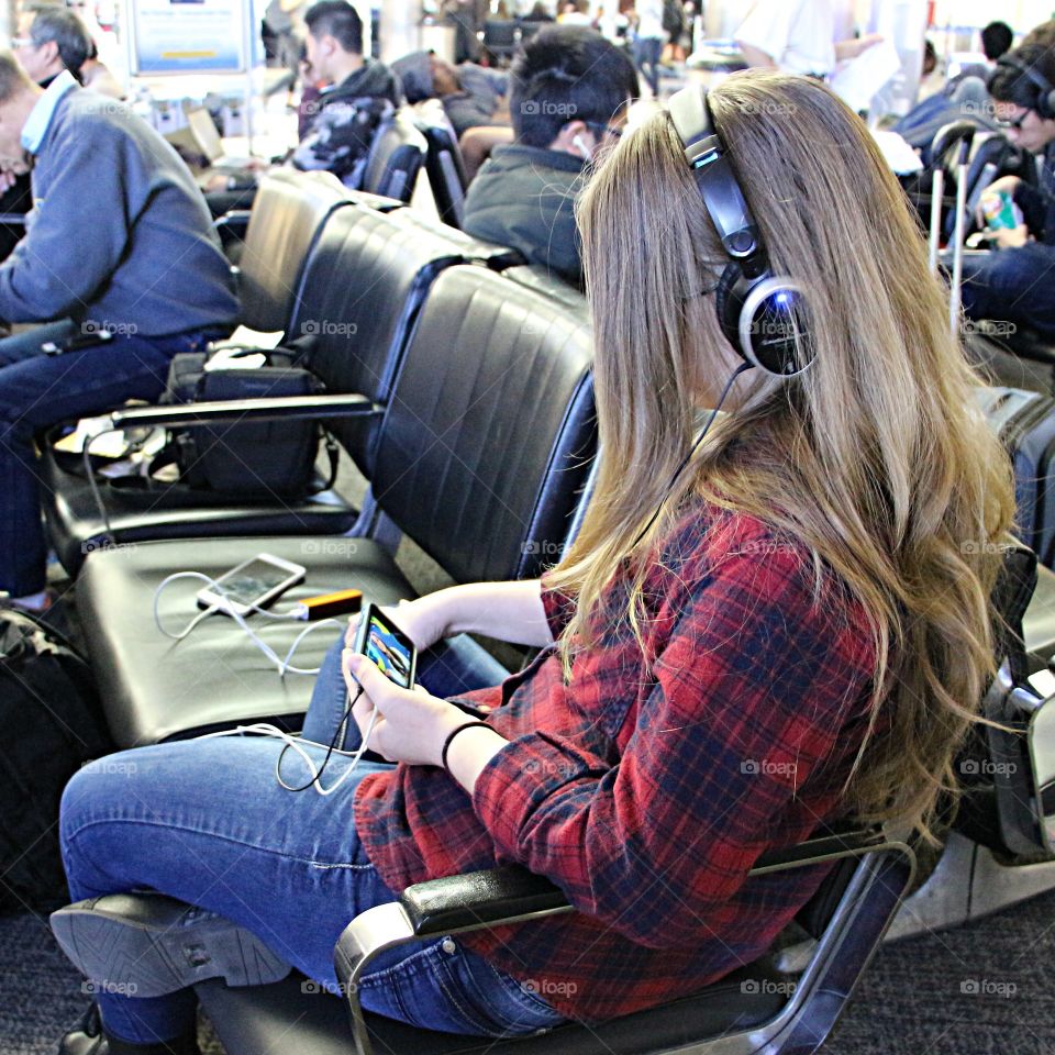 A girl with her headphone is listening to music from her cellphone while waiting for boarding at the airport
