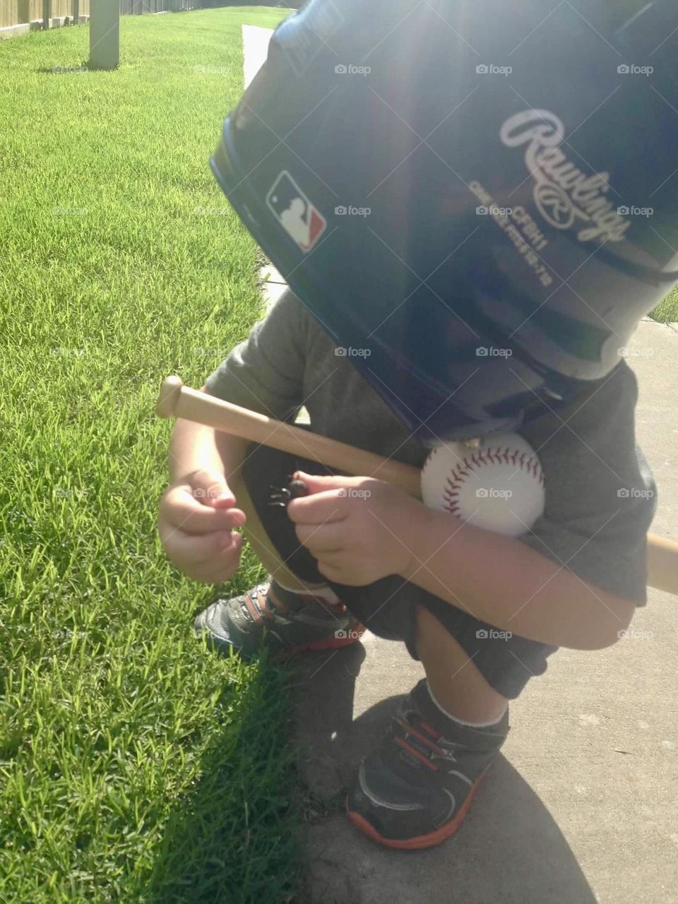 A toddler boy stands outside on the green grass and sidewalk holds his baseball, bat, and glove while wearing his “helmet hat.”