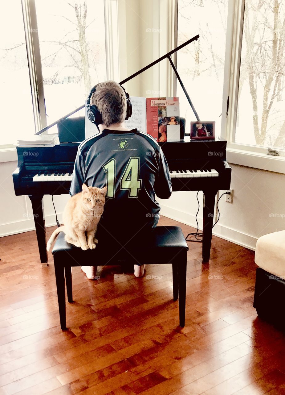 Young man sitting playing piano which is one of his favorite hobbies while his orange tabby is sitting right by him while he plays! 