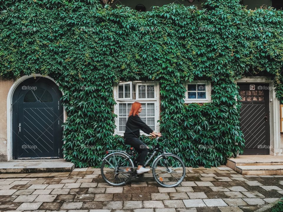 Woman riding bicycle in front of house with vine