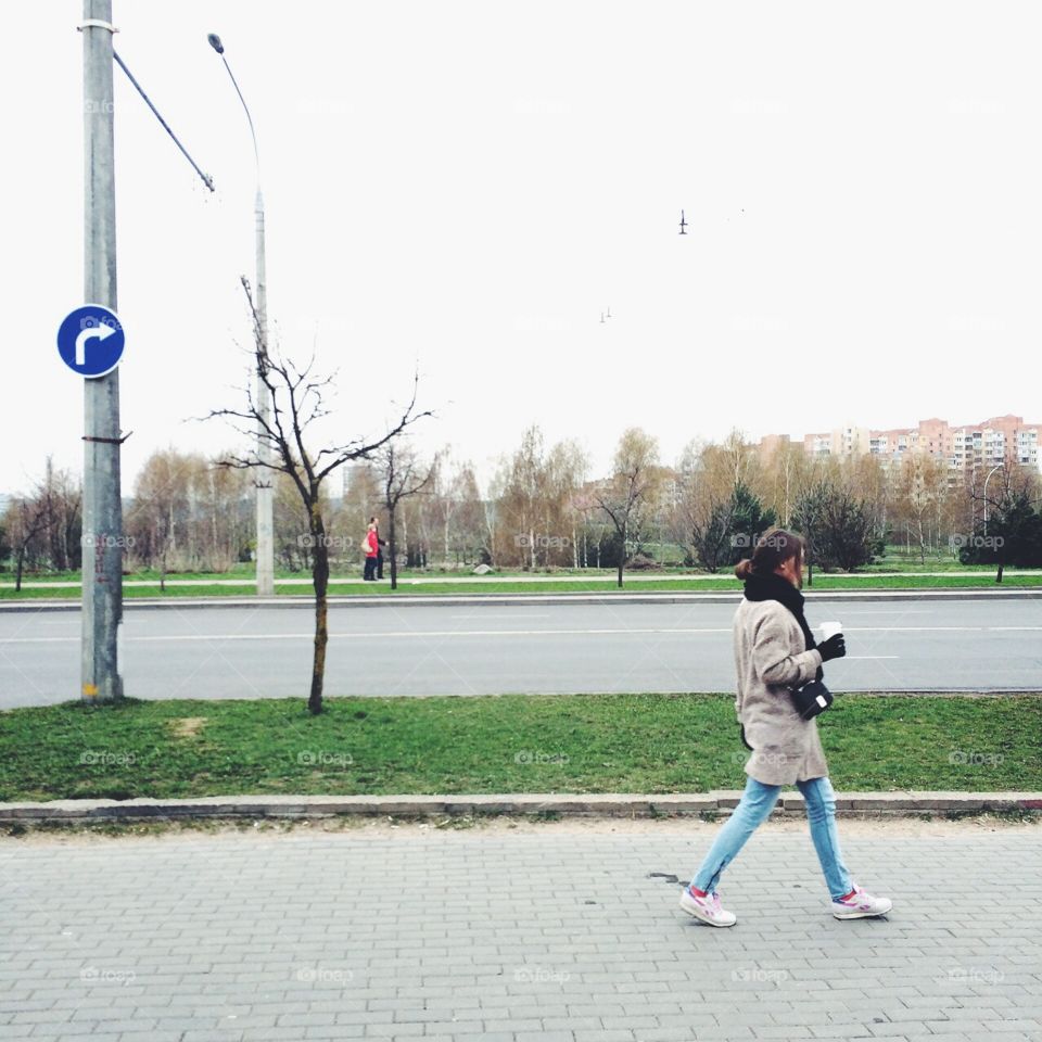 A brunette girl, wearing a grey coat, sneakers, big knitted scarf, blue jeans and a crossbody bag, walks down the autumn empty city street