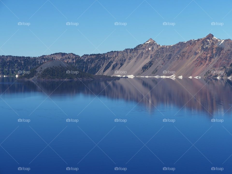 Wizard Island in bright blue Crater Lake that still has a little ice on it and snow on the jagged rim reflecting in the water on a sunny summer morning in Southern Oregon. 