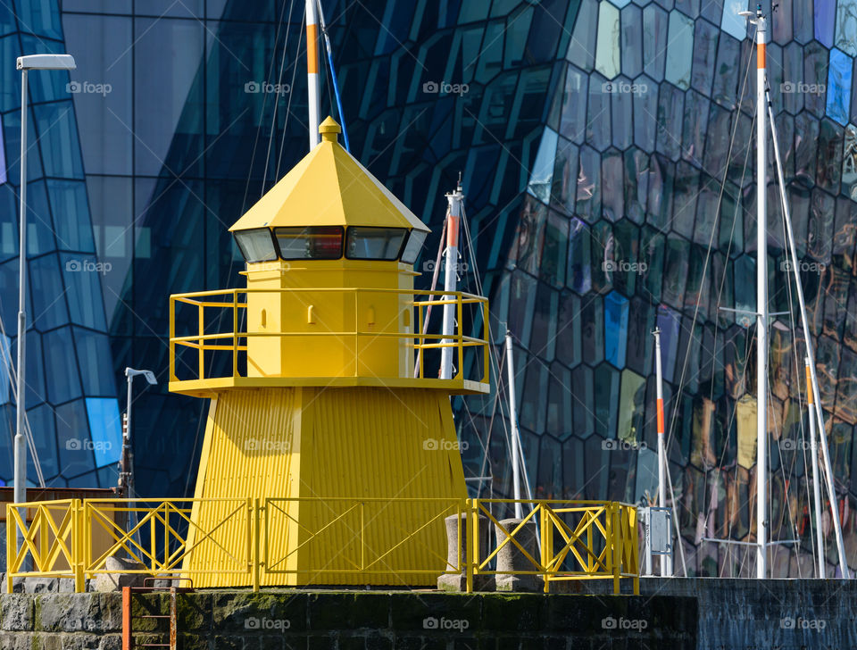 REYKJAVIK, ICELAND - JUNE 3, 2017: Yellow lighthouse in the Reykjavik, Iceland harbor with colorful and abstract glass exterior of the Harpa concert on the background with reflections in the windows and masts of the sailing boats in between.