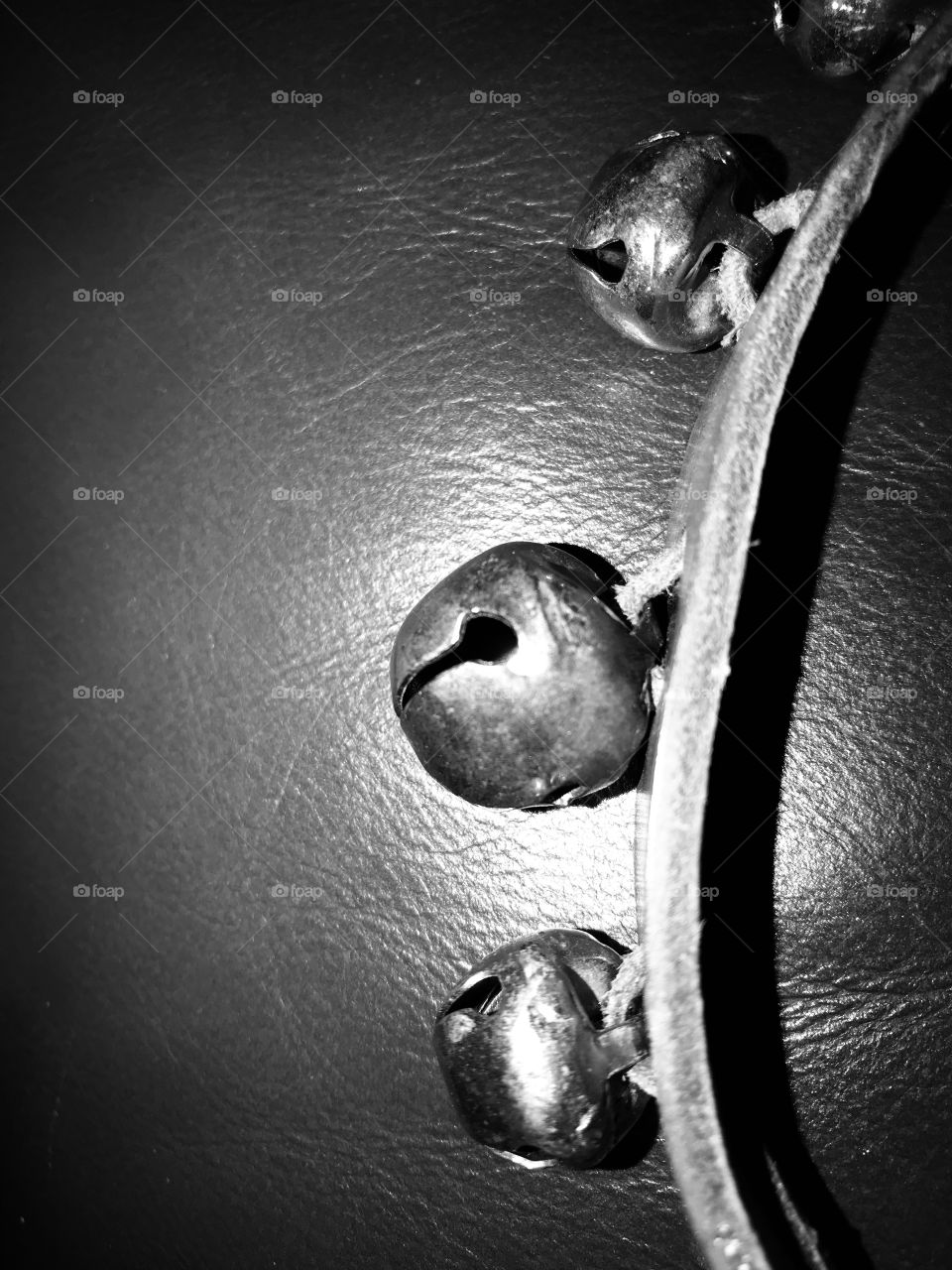 Black & white closeup of a set of handmade Christmas jingle bells made of wood, leather, leather rope ties and round silvery bells. The bells are sitting on the arm of a chair and are lit by a variety of Christmas lights throughout the room.