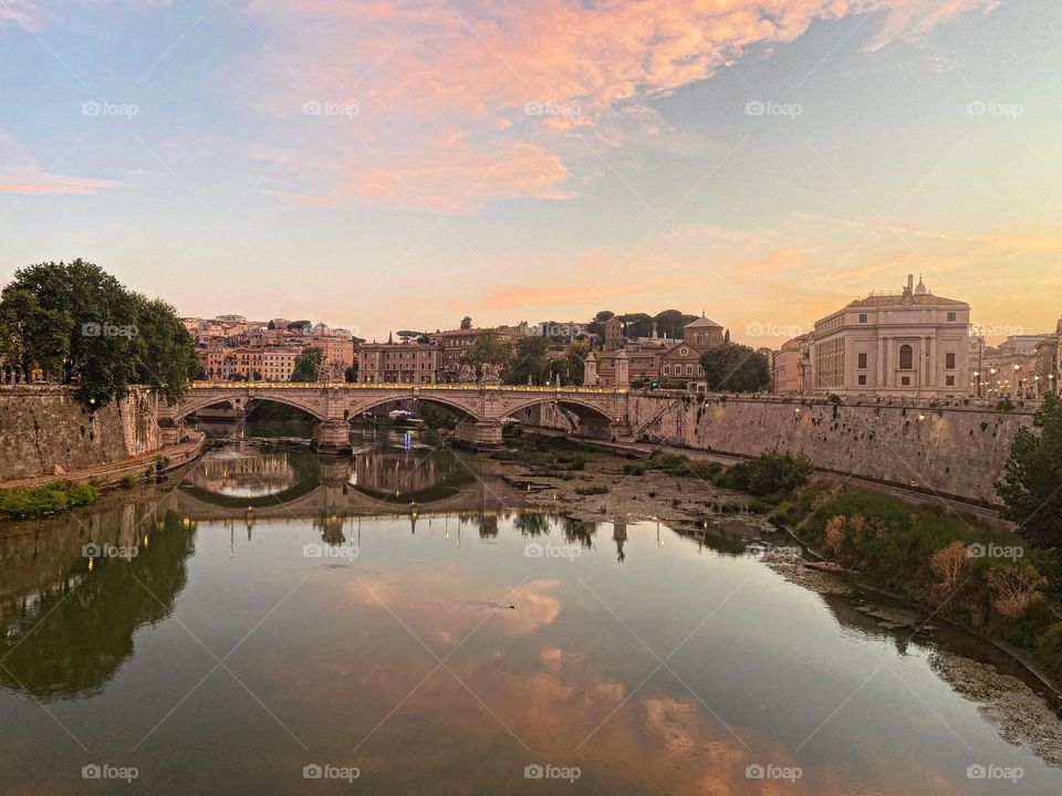 The Tiber River smoothly reflects the cotton candy sky with an arched bridge and ancient Roman buildings 