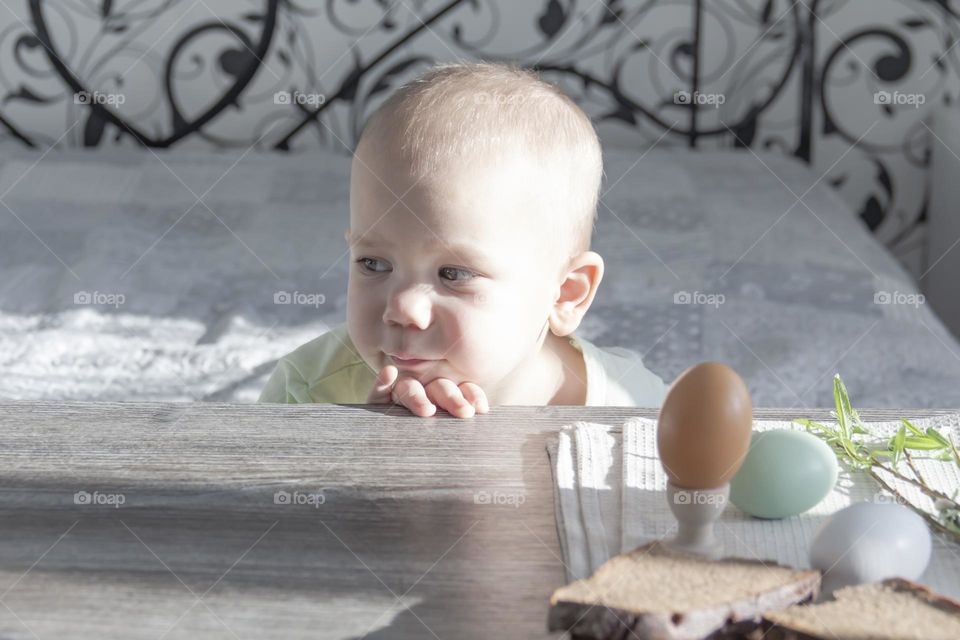A little boy with a thoughtful and sad face sits at a wooden, gray table with bread and Easter eggs on it.