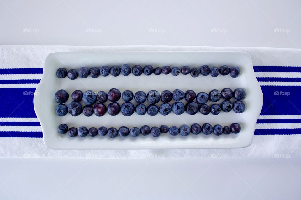 Horizontal flat lay of blueberries on a rectangular white plate, arranged in lines to emphasize the blue stripes on the white dish towel underneath, all on a white background 