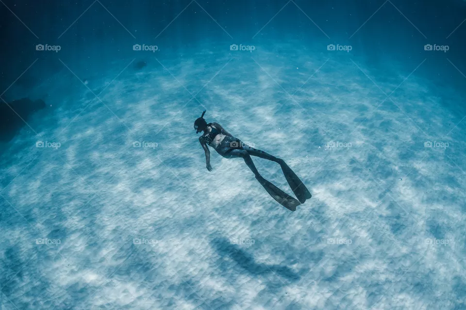 A female free diver swims above the white sand in crystal clear turquoise ocean water.