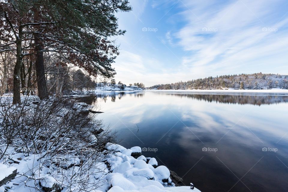 Beautiful winter landscape, snow covered trees by a peaceful lake with reflections from the blue sky and clouds 