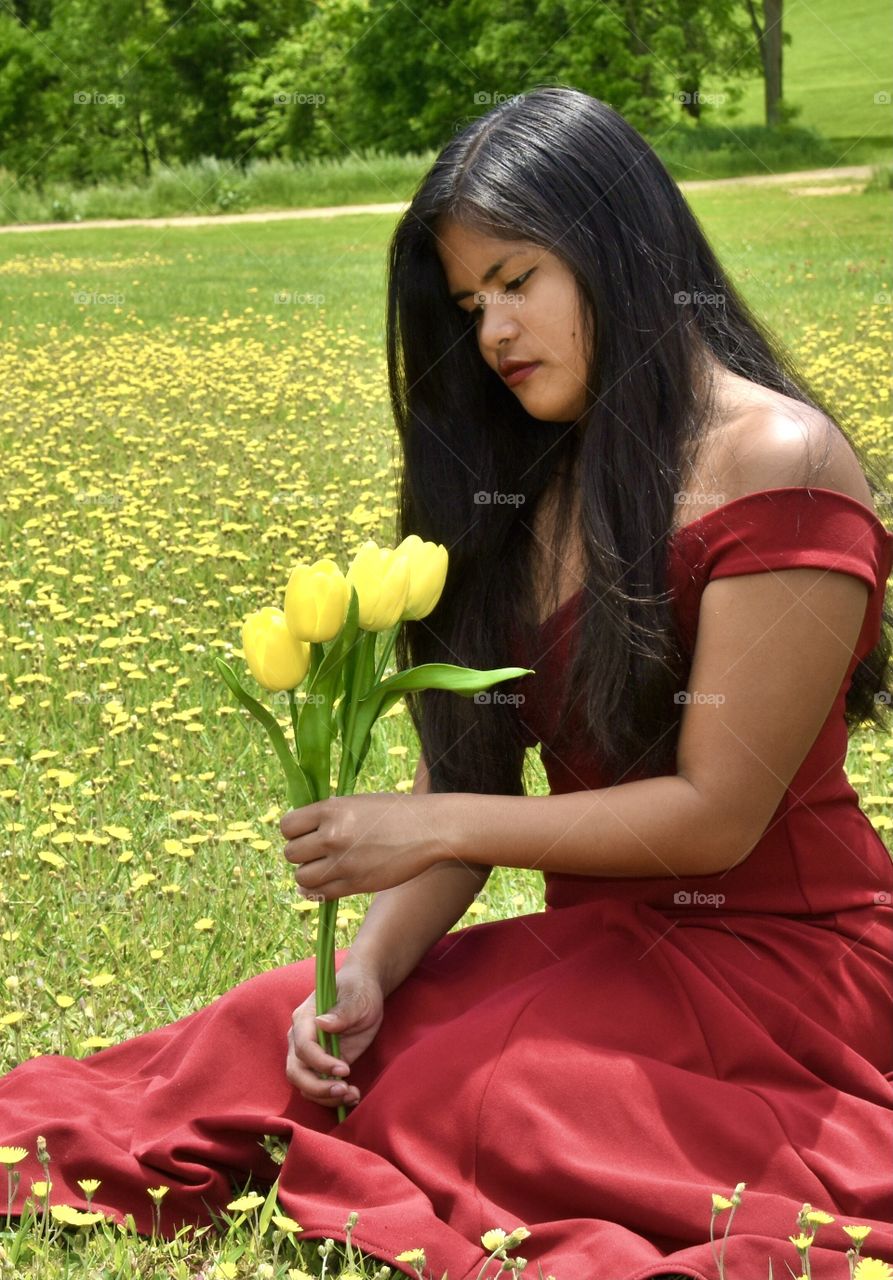 Young lady in a red gown sitting in a field of yellow flowers and holding yellow tulips 