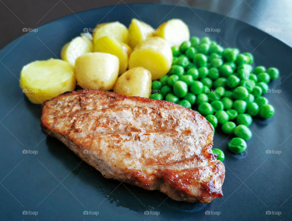 Grilled pork chop or pork steak with baby potatoes and peas.  Selective focus at the piece of pork chop and copy space is at the blurry part of  the photo.