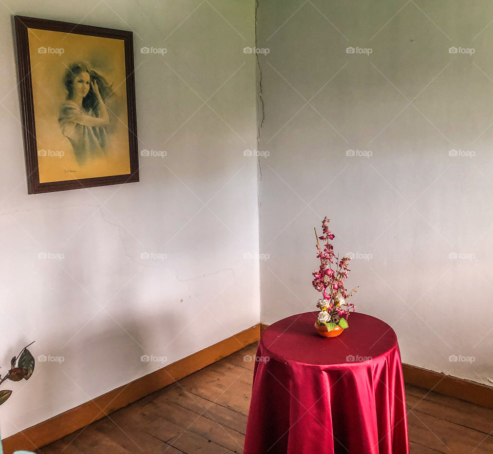 An abandoned room, with wooden floors and cracks in the walls. A large image of a girl combing her hair is on a wall and a small table draped in a bright red cloth still in place